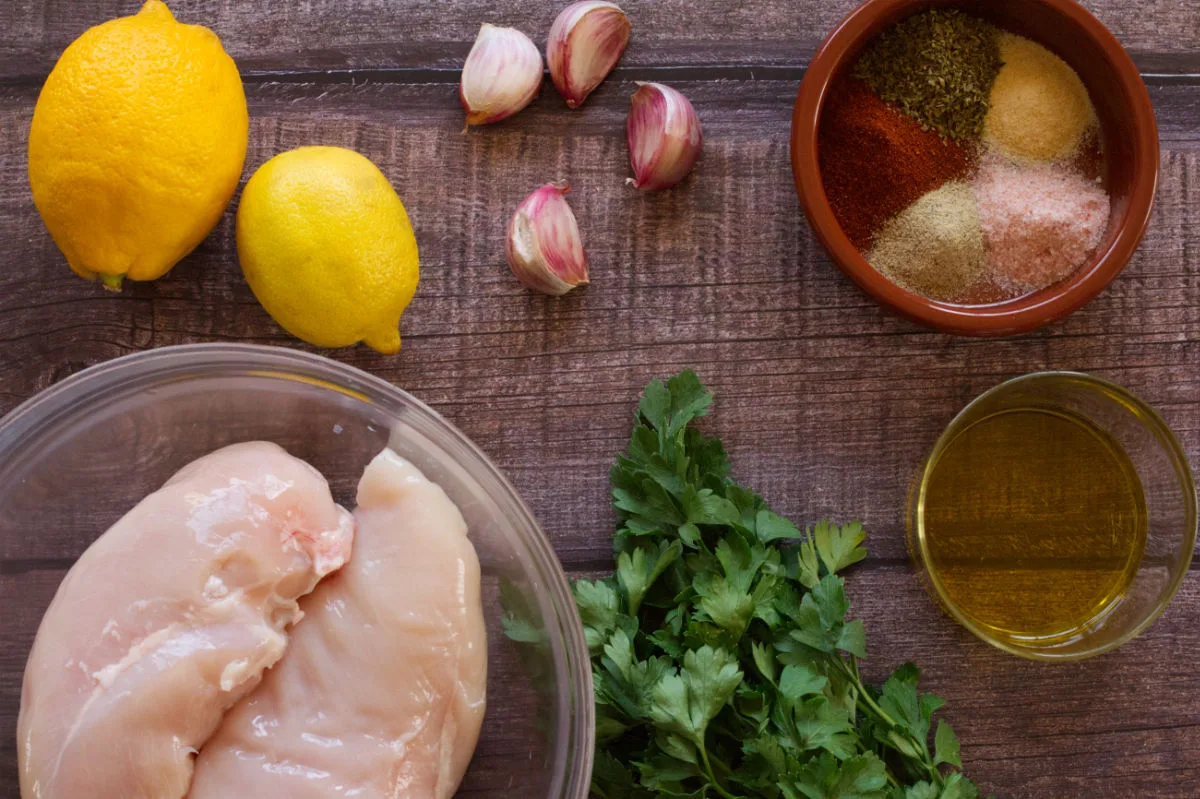 A bowl of uncooked chicken breasts sits beside some fresh lemons, garlic, spices, and olive oil. 