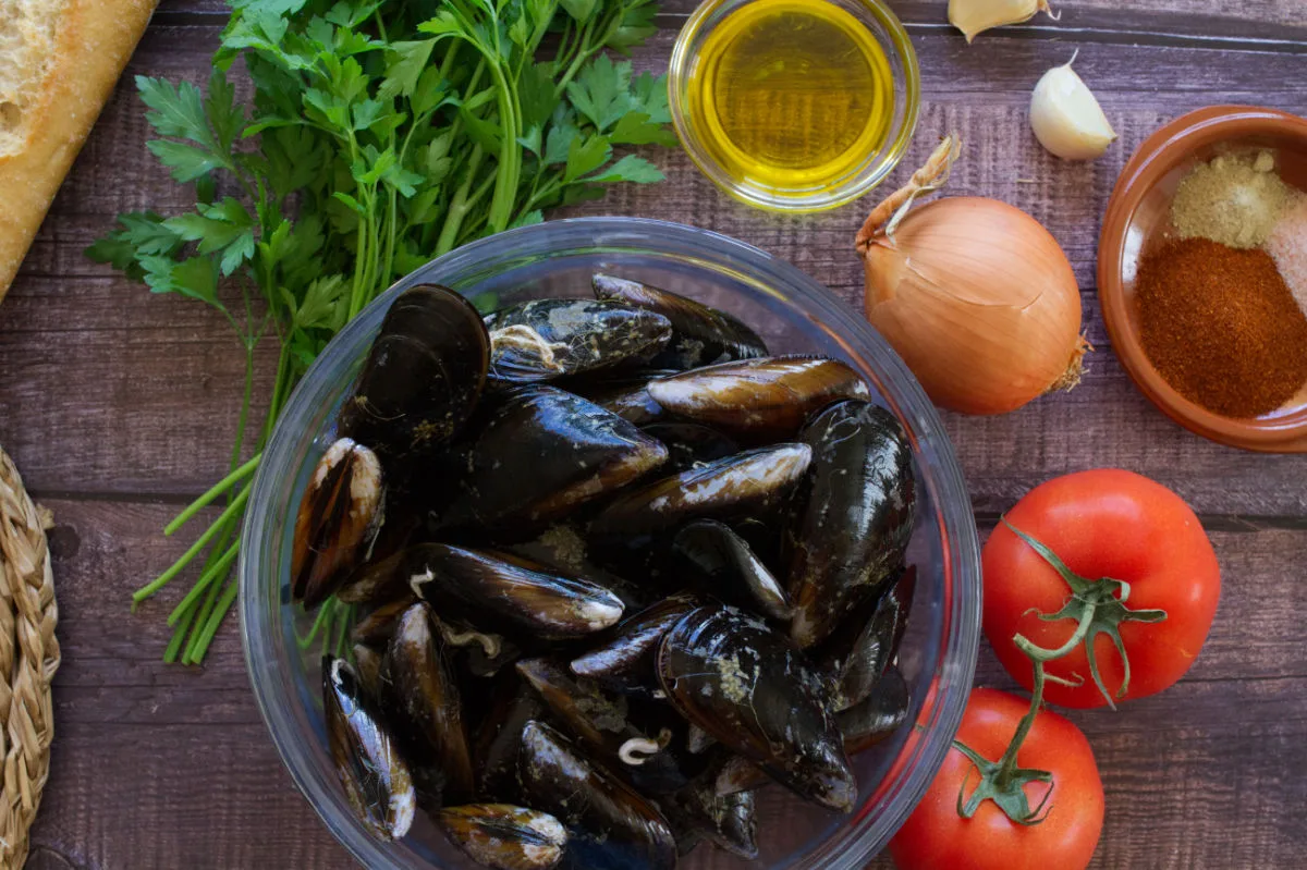 A bowl of uncooked mussels sits next to some tomatoes, onion, garlic, spices, and oil. 