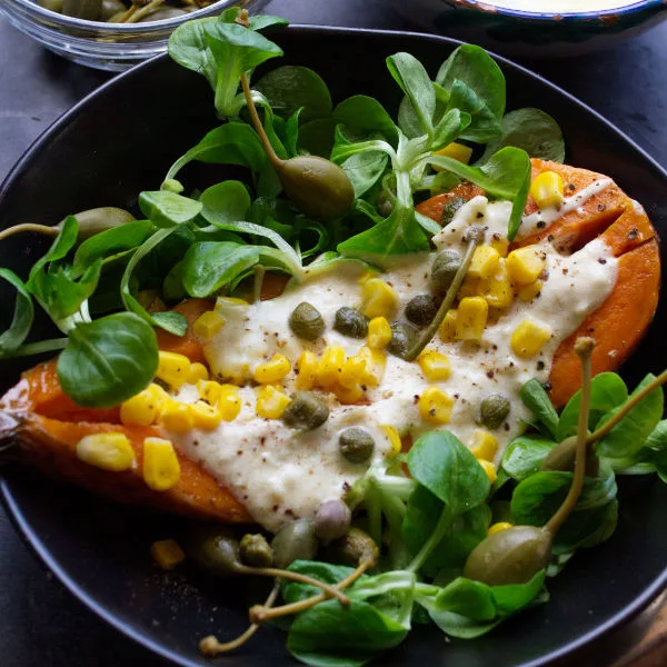 A bowl of baked sweet potato with toppings, salad, and a yogurt dressing.