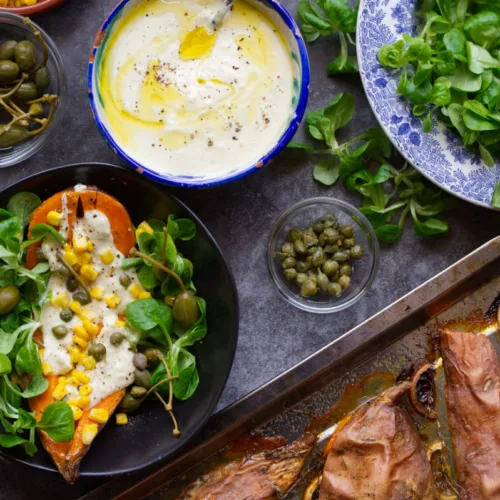 A bowl of baked sweet potato sits beside a tangy yogurt dressing and sheet pan of sweet potatoes