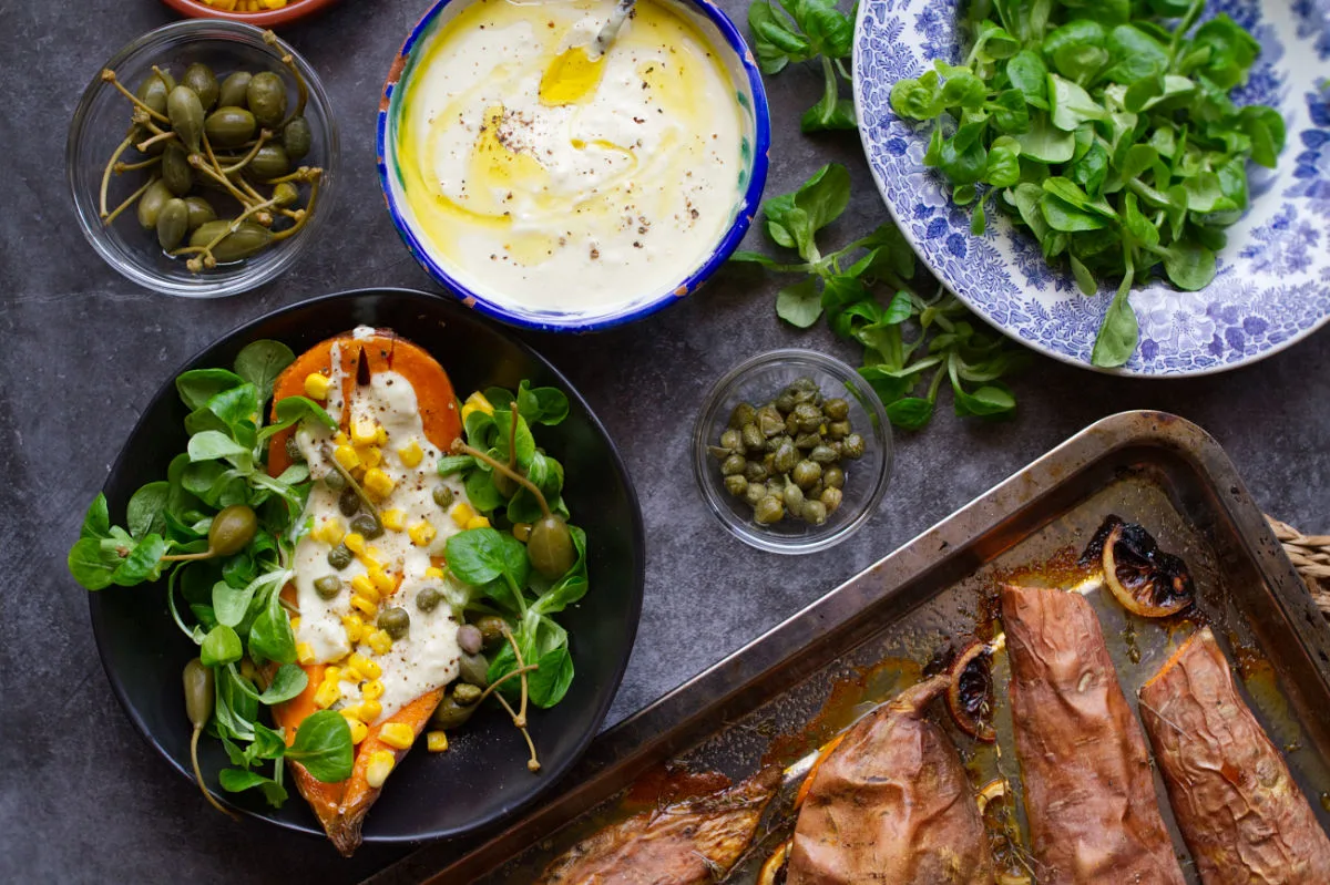 A bowl of baked sweet potato sits beside a tangy yogurt dressing and sheet pan of sweet potatoes