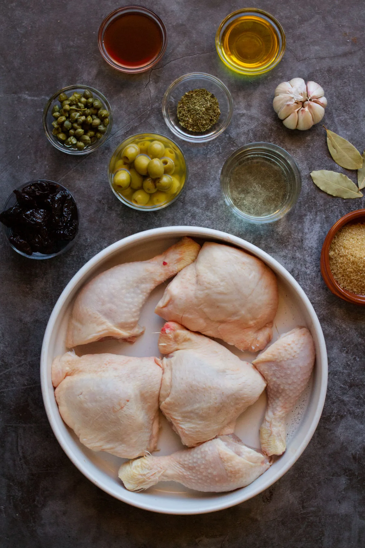 A large casserole dish loaded with chicken pieces sits besides small bowls of spices and ingredients used to make chicken marbella.