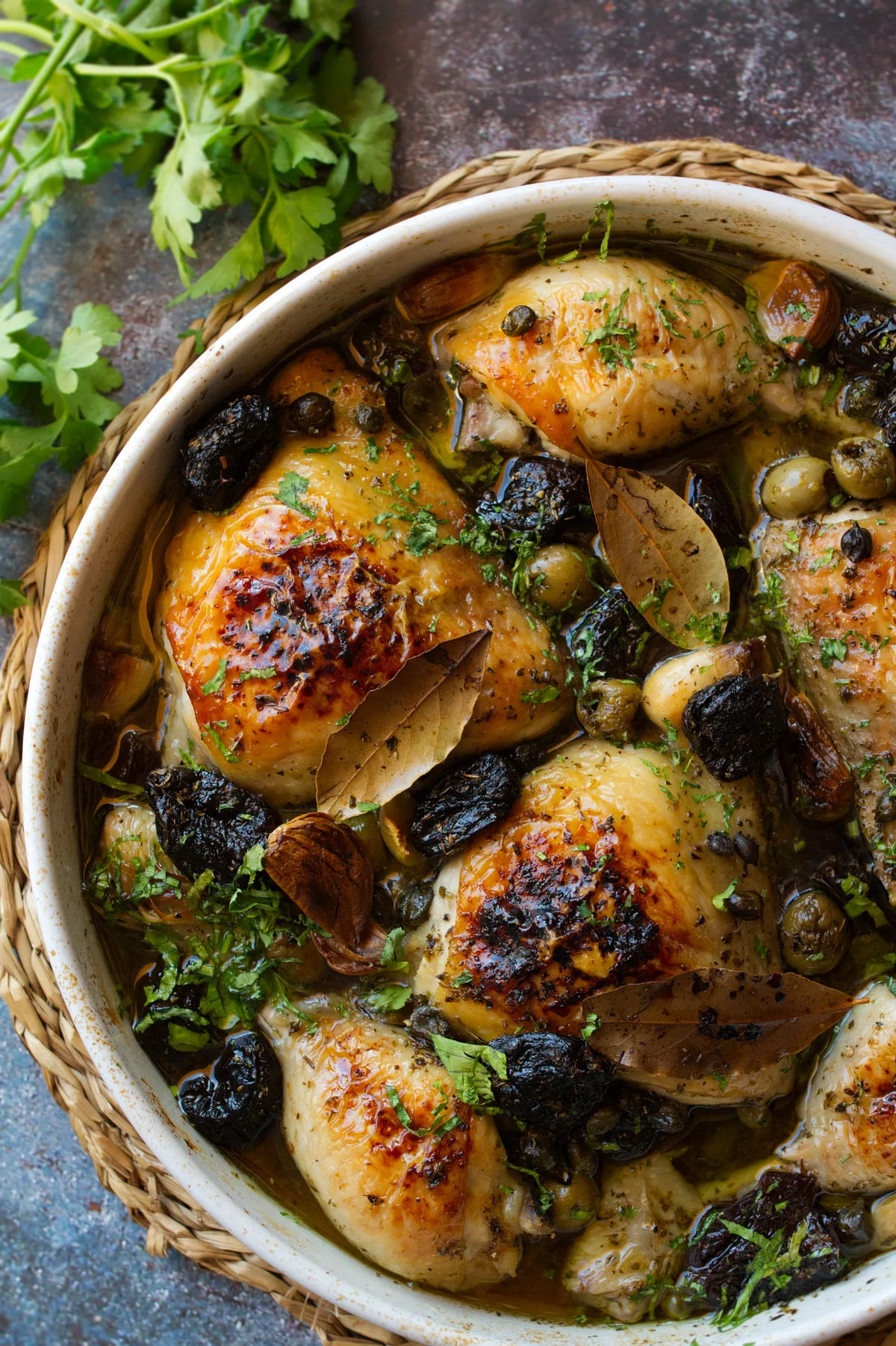 A large casserole dish of chicken Marbella sist beside some fresh herbs and lemon.