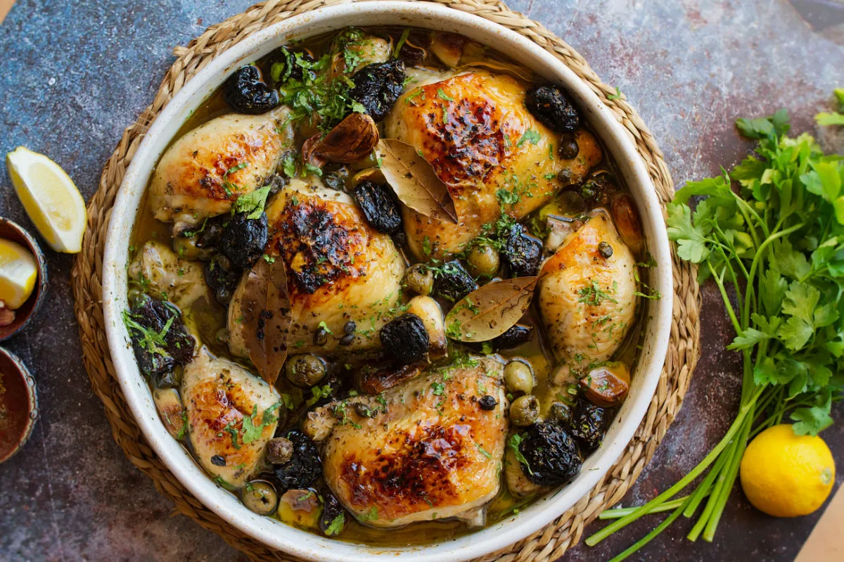 A large casserole dish of chicken Marbella sist beside some fresh herbs and lemon.