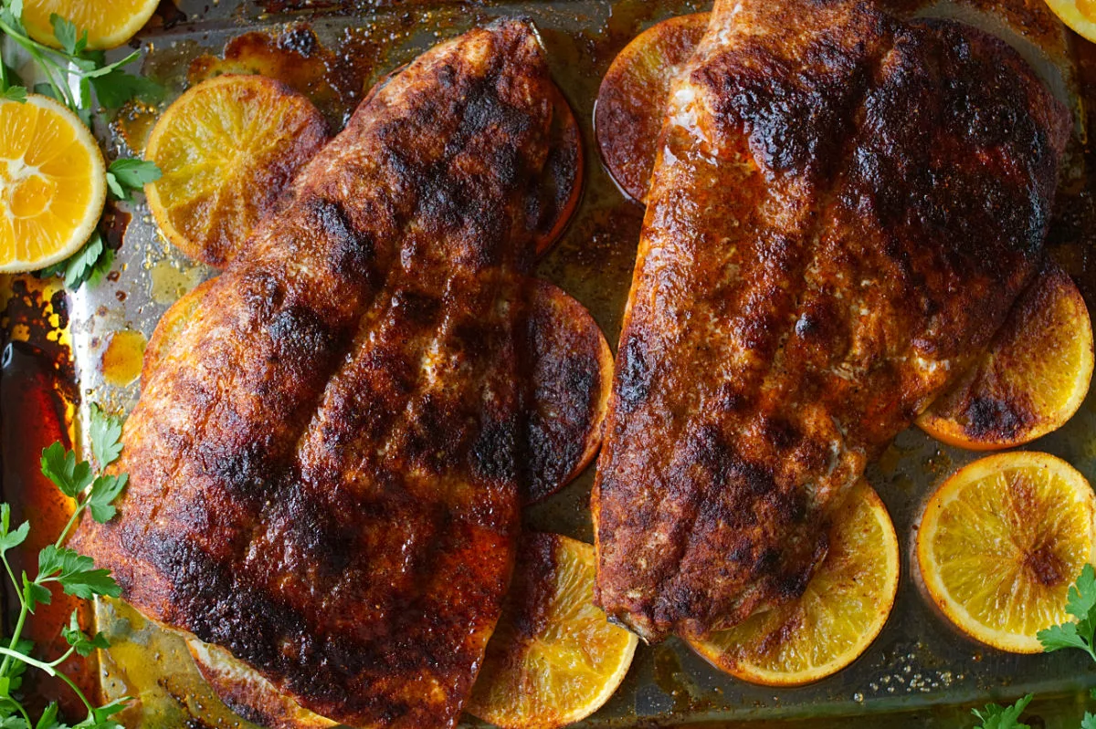 A large fillet of Spanish-style spicy salmon beside some orange rounds and fresh herbs.