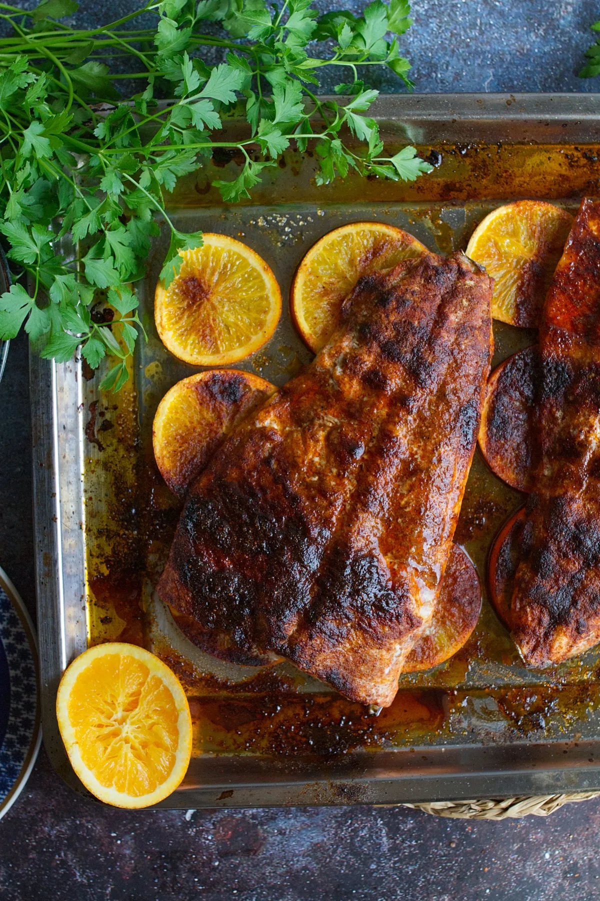 A large fillet of Spanish-style spicy salmon beside some orange rounds and fresh herbs.