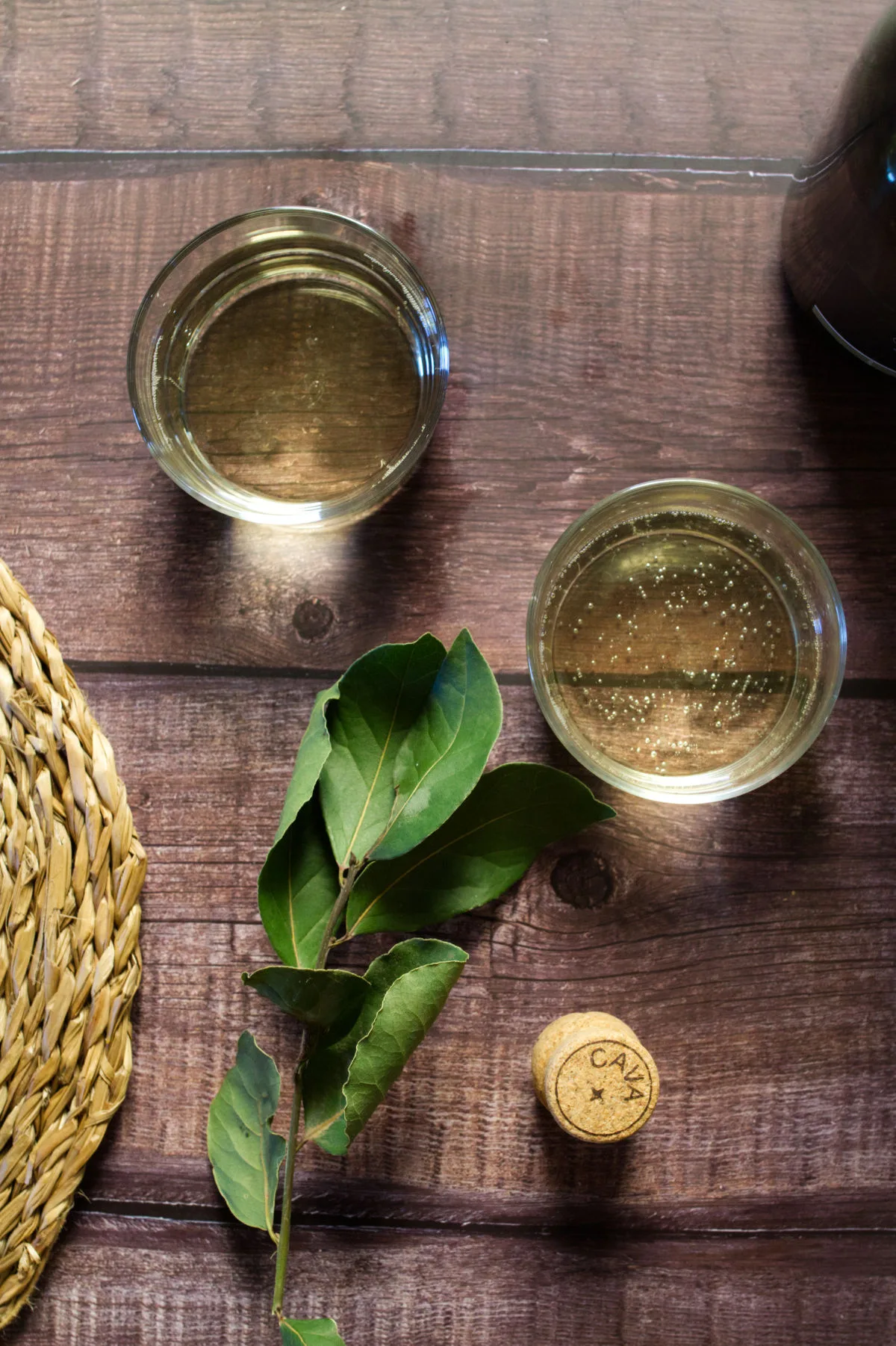 A cava bottle cork sits beside two glasses of cava and some bay leaves. 