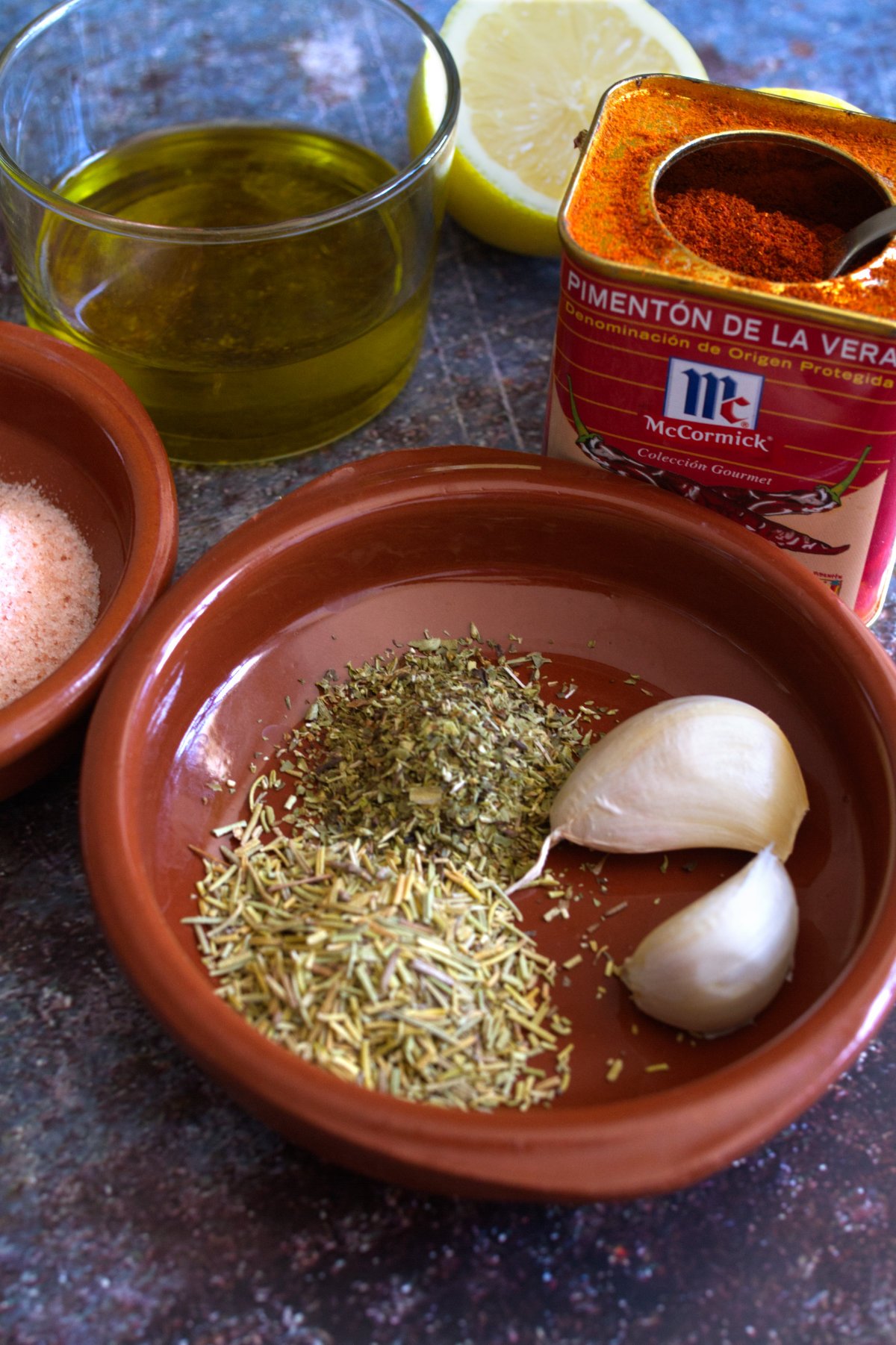 Spanish paprika sits beside some herbs, garlic cloves, and fresh lemons.